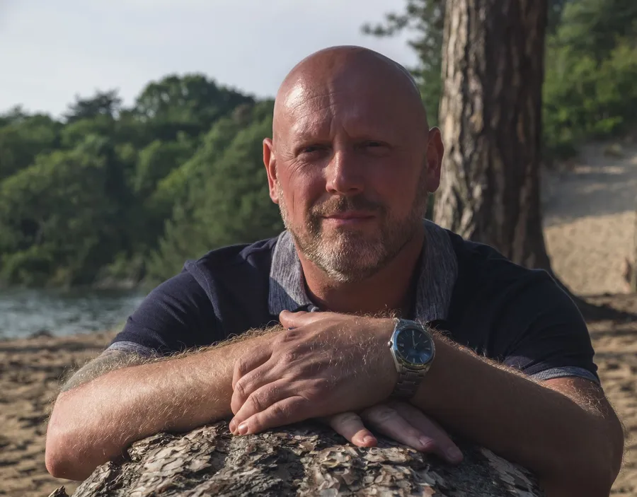 Patrick leunt met zijn armen op een boomstam bij het water en kijkt in de camera