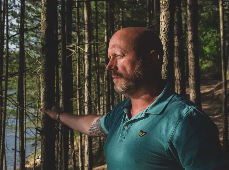 Patrick staat tussen de bomen in het bos, hand tegen een stam, en kijkt in de verte